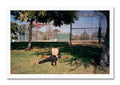 Person lying on grass with a paper bag over their head in an outdoor setting with trees and a fence.